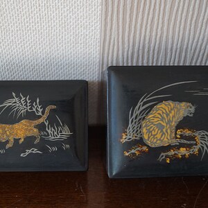 May include: Two black lacquered boxes with gold and silver designs. One box features a tiger in a grassy field with a moon in the background. The other box features a leopard running through a grassy field.
