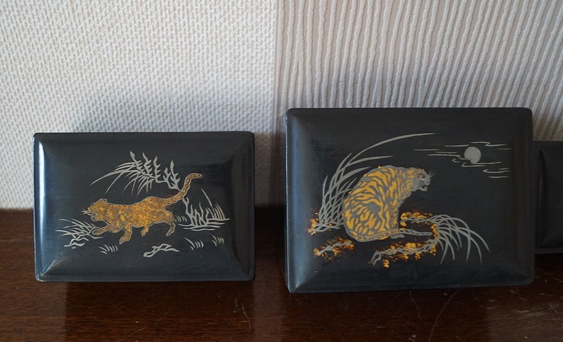 May include: Two black lacquered boxes with gold and silver designs. One box features a tiger in a grassy field with a moon in the background. The other box features a leopard running through a grassy field.