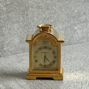 May include: A gold-toned Wellington quartz mantel clock with a metallic finish, featuring a square face with Roman numerals, a decorative embossed pattern, and a small handle on the top. The clock displays the text Wellington and Tempus Fugit.