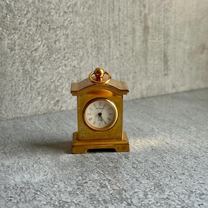May include: A small gold-toned mantel clock with a rectangular base and a rounded top featuring a circular handle. The clock face is white with black Roman numerals and the text Wellington printed in the centre, featuring black hour and minute hands.