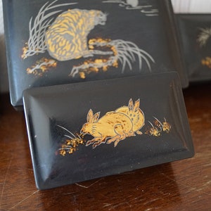 May include: Four black lacquered boxes with gold painted designs. The boxes feature a tiger, a rabbit, a bird, and a fish. The boxes are arranged in a row on a wooden surface.