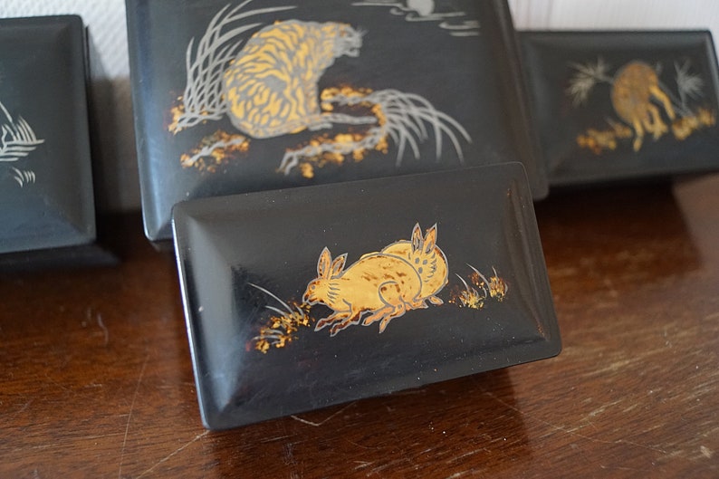 May include: Four black lacquered boxes with gold painted designs. The boxes feature a tiger, a rabbit, a bird, and a fish. The boxes are arranged in a row on a wooden surface.