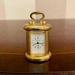 Peut inclure: Une petite horloge à quartz dorée avec un cadran rectangulaire et des chiffres romains. L'horloge a une poignée en haut et le mot "Wellington" est imprimé sur le cadran. L'horloge mesure environ 7.6 cm de haut.