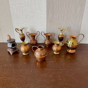 May include: A selection of miniature metal jugs and a small stove. The jugs are in various shapes, sizes, and colours, with copper and brass hues. Some have handles and decorative patterns. The stove is silver and brown.
