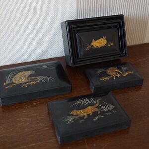 May include: Set of four black lacquered boxes with gold and silver animal designs. The boxes are nested inside each other and feature a tiger, a rabbit, a cat, and a deer.