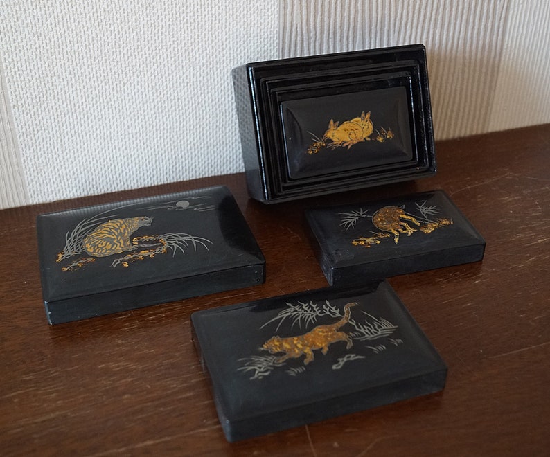 May include: Set of four black lacquered boxes with gold and silver animal designs. The boxes are nested inside each other and feature a tiger, a rabbit, a cat, and a deer.