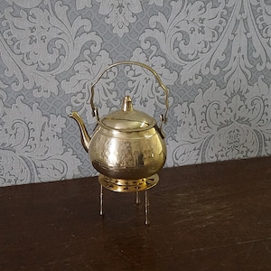 Könnte beinhalten: Eine kleine, goldfarbene Messing-Teekanne mit einem verzierten Design steht auf einem dreibeinigen Ständer. Die Teekanne hat einen Griff und einen Ausguss.