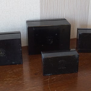 May include: Four black square boxes with a raised design on the top. The boxes are arranged in a row, with the largest box in the middle and the smallest box on the right.