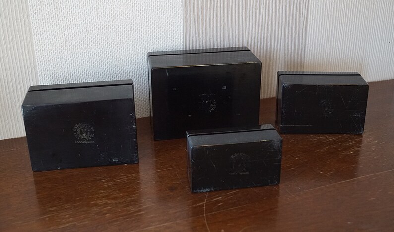 May include: Four black square boxes with a raised design on the top. The boxes are arranged in a row, with the largest box in the middle and the smallest box on the right.