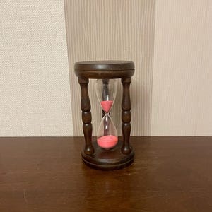 Op de afbeelding: Een houten zandloper met een roze zandtimer. De zandloper staat op een houten oppervlak.