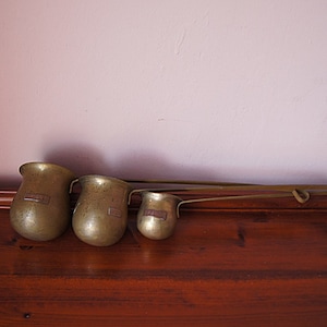 May include: Three brass measuring cups with long handles, the largest is about 15 cm tall, the smallest is about 7.5 cm tall, they are on a wooden surface.