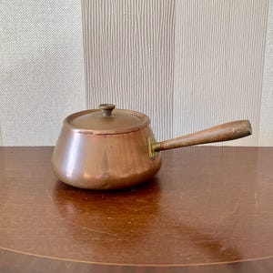 May include: A small copper saucepan with a wooden handle and a lid. The saucepan has a brass accent on the handle.