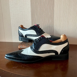 以下が含まれることがあります： クラシックなデザインの、黒と白のウィングチップドレスシューズのペア。靴はパーフォレーションパターンと赤い内装が特徴です。靴は木製の表面に展示されています。