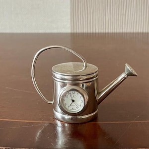 Op de afbeelding: Een miniatuur zilverkleurige metalen gieter met een geïntegreerde analoge kwartsklok aan de zijkant. De wijzerplaat is wit met zwarte cijfers en wijzers, en de gieter heeft een gebogen handvat en een kleine tuit.