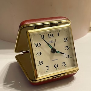 Op de afbeelding: Een vintage rode en gouden reiswekker. De wijzerplaat is crèmekleurig met zwarte cijfers en wijzers, en het woord "Atlanta" staat op de wijzerplaat. De klok zit in een rode behuizing die open en dicht kan.
