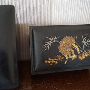 May include: Two black wooden boxes with intricate designs. The box on the right has a gold and silver design of a deer in a bamboo forest.