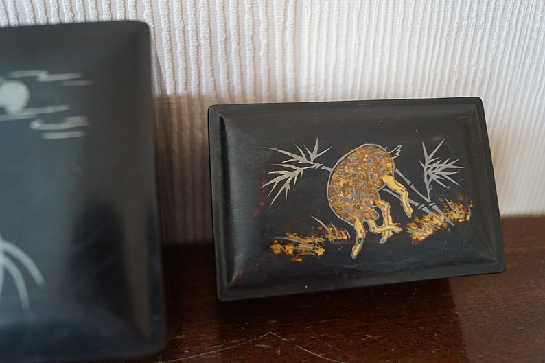 May include: Two black wooden boxes with intricate designs. The box on the right has a gold and silver design of a deer in a bamboo forest.