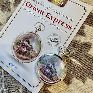 May include: Two silver-plated pocket watches with a train scene inside, labeled "Orient Express." The watches are on a certificate of authenticity with the text "Glory of Steam." The watches are silver-plated and guaranteed.