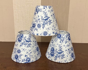 Puede incluir: Tres pantallas de lámpara cónicas con fondo blanco, un patrón floral azul e ilustraciones de pájaros pequeños. Las pantallas están dispuestas en formación triangular con una pantalla colocada sobre las otras dos.