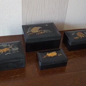 May include: Four black lacquered boxes with gold and silver animal designs. The boxes are stacked in a pyramid shape on a wooden surface.
