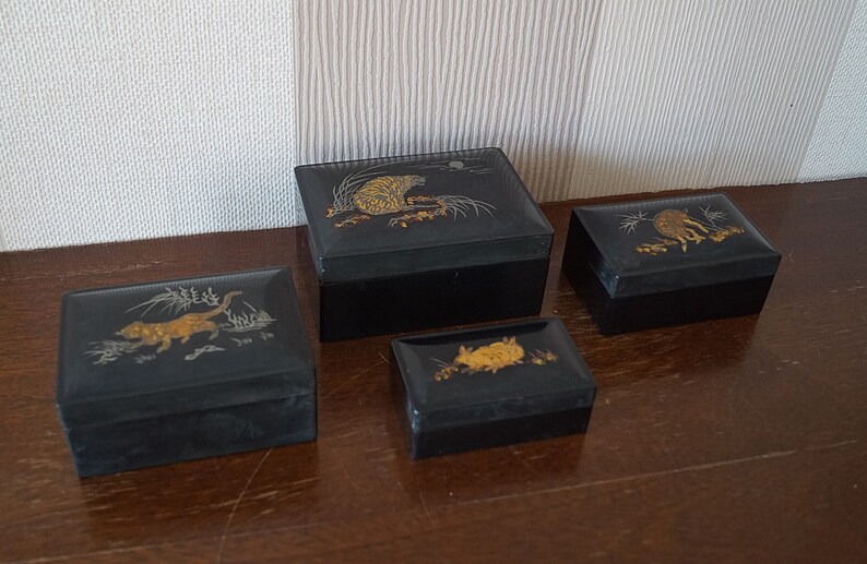 May include: Four black lacquered boxes with gold and silver animal designs. The boxes are stacked in a pyramid shape on a wooden surface.