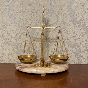 May include: A classic brass balance scale with two circular bowls hanging from chains. The scale is set on a marble base and includes a set of brass weights. The background features patterned wallpaper.