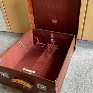 May include: An open, vintage brown suitcase with a red interior. The suitcase has metal latches and a leather handle. The interior features a red fabric lining and several metal hooks. The suitcase is likely used for travel and storage.