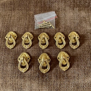 May include: Eight ornate, gold-coloured drawer pulls with a ring handle and decorative detailing. A small bag of screws is also visible. The pulls are arranged on a brown, textured surface, suggesting a hardware or home decor theme.