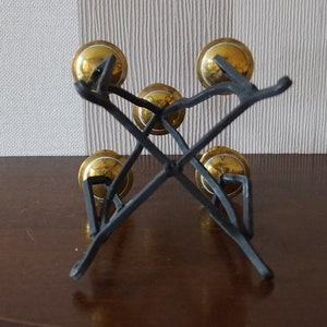 May include: A set of five gold bells hanging from a black metal stand. The bells are arranged in a circular pattern and are suspended from the stand by a series of black metal rods.