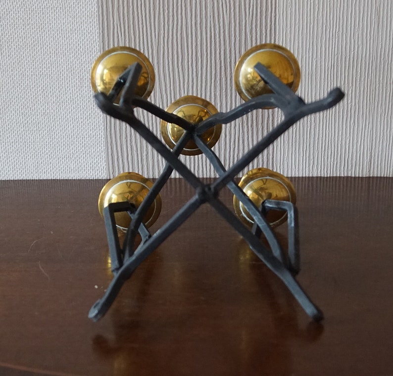 May include: A set of five gold bells hanging from a black metal stand. The bells are arranged in a circular pattern and are suspended from the stand by a series of black metal rods.