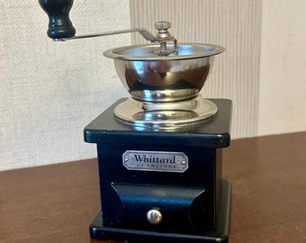 May include: A manual coffee grinder with a black wooden square base, a silver metal hopper, and a black handle knob. A silver metal plate on the front features the text Whittard of Chelsea. A small silver drawer knob is centered below the plate.