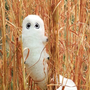 Könnte beinhalten: Zwei weiße Plüschtiere mit großen schwarzen Augen und kleinen schwarzen Füßen sind zwischen hohem, trockenem Gras versteckt. Die Spielzeuge haben eine abgerundete, wurmartige Form und scheinen handgefertigt zu sein. Der Hintergrund zeigt eine Mischung aus grüner und brauner Vegetation.