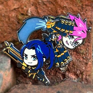 May include: Enamel pin depicting two cartoon characters with blue and pink hair, wielding weapons. They are dressed in black and gold attire. The pin is set against a textured, reddish-brown background.