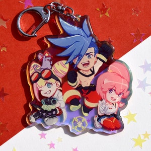 May include: A colorful anime-style keychain featuring three characters: a blue-haired boy in a red and black outfit, a blonde girl with goggles and a yellow outfit, and a pink-haired girl in a red and white outfit. The keychain is made of clear acrylic with a holographic finish.