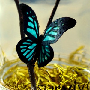 May include: A decorative butterfly with black wings and turquoise accents, perched on a thin brown branch. The butterfly's wings have intricate cut-out patterns. The background is blurred, with hints of yellow and a light-colored sky.