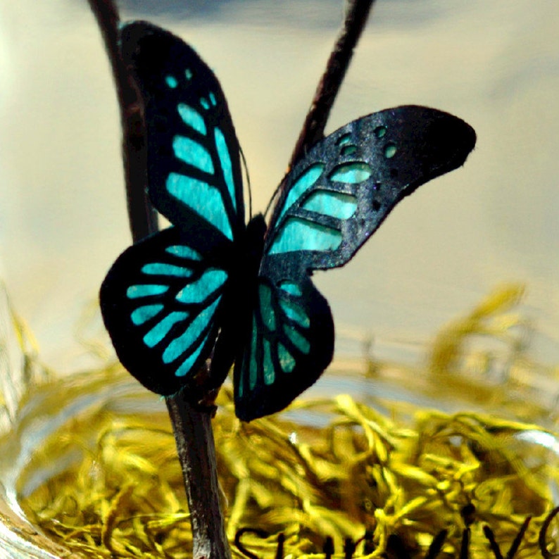 May include: A decorative butterfly with black wings and turquoise accents, perched on a thin brown branch. The butterfly's wings have intricate cut-out patterns. The background is blurred, with hints of yellow and a light-colored sky.