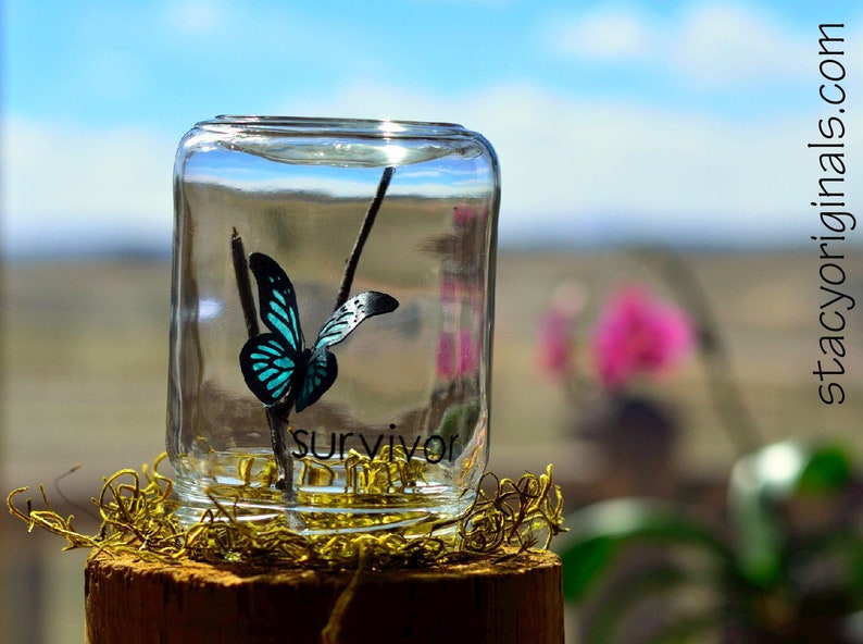 May include: A clear glass jar with a teal and black butterfly inside, perched on a twig. The word "SURVIVOR" is printed on the jar. The jar sits on a wooden base with decorative gold moss.