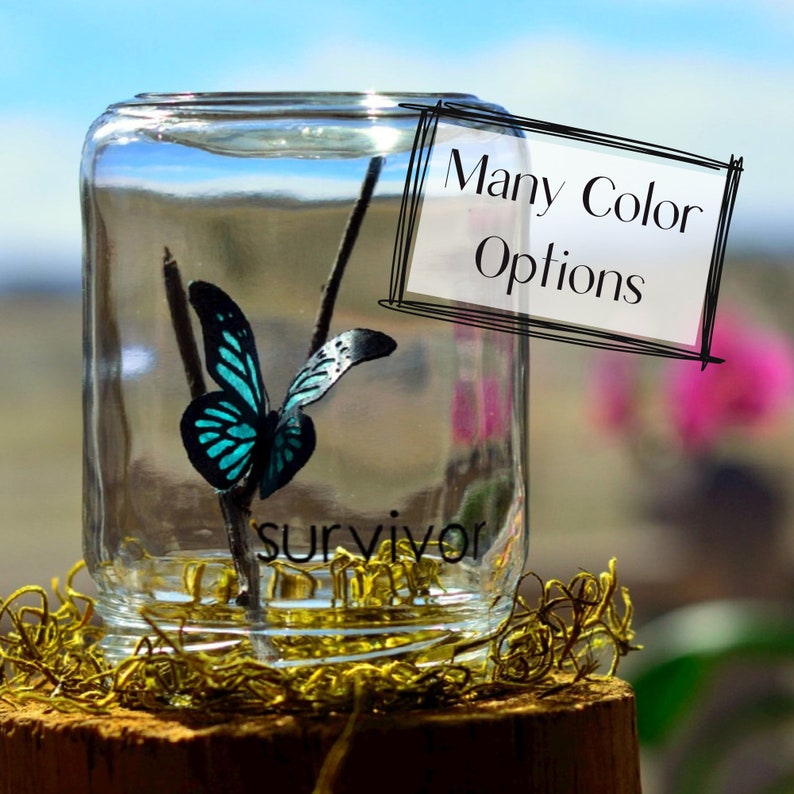 May include: A clear glass jar with a teal and black butterfly inside, perched on a twig. The word "survivor" is at the bottom. A sign reads "Many Color Options". The jar sits on a wooden base with gold decorative moss.
