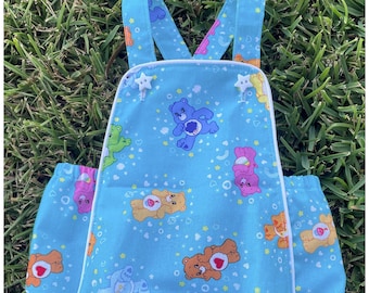 May include: A light blue baby onesie with white straps and white buttons. The onesie has a print of Care Bears on a blue background with white stars.