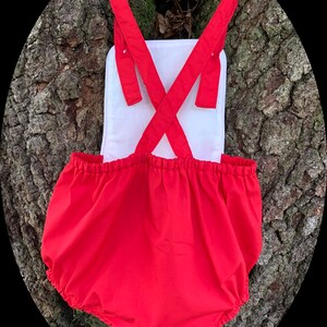 May include: Red baby bloomers with white straps and a white back panel. The bloomers have a gathered elastic waistband and are made of a soft, comfortable fabric.