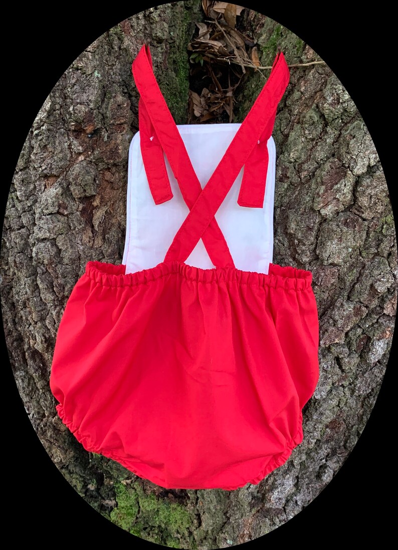 May include: Red baby bloomers with white straps and a white back panel. The bloomers have a gathered elastic waistband and are made of a soft, comfortable fabric.