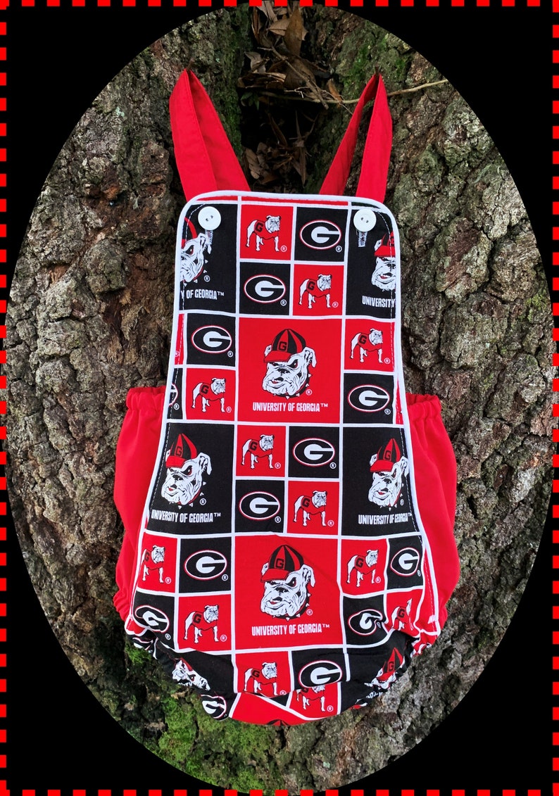 May include: Red and black baby onesie with a white trim. The onesie has a checkered pattern with the University of Georgia logo and a bulldog mascot.
