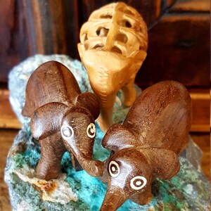 May include: Three wooden elephant figurines, two brown and one light brown, are arranged on a turquoise and gray rock. The elephants have white painted eyes and are carved in a simple style.