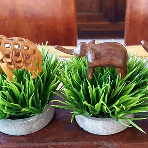 May include: Three wooden elephant figurines, each standing on a bed of green artificial grass in a gray concrete pot. The elephants are carved in different styles, with one being a more intricate design.
