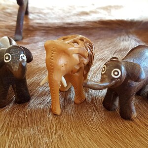 May include: Three wooden elephant figurines, one brown, one light brown, and one dark brown. The figurines are standing on a brown and white cowhide rug.