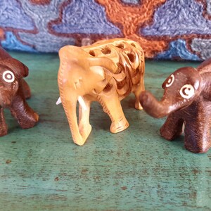 May include: Three wooden elephant figurines, one is light brown with intricate carvings, the other two are dark brown with white painted eyes.
