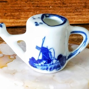 May include: A small white ceramic watering can with a blue and white windmill design. The watering can has a spout and a handle.