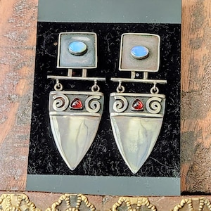 May include: Silver-toned earrings with a geometric design. Each earring features a square top with a blue stone, a horizontal bar, and a shield-shaped bottom with a red stone and spiral accents. The earrings are displayed on a black surface.