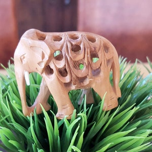 May include: A small, intricately carved wooden elephant figurine with a light brown color. The elephant is standing on a bed of green artificial grass.