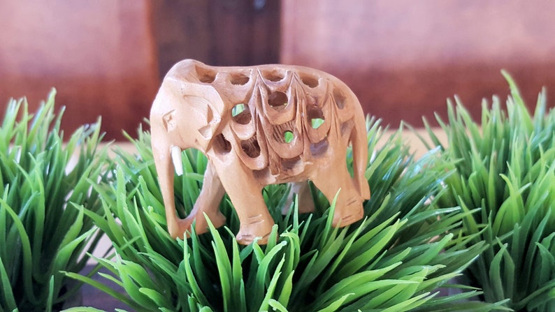 May include: A small, intricately carved wooden elephant figurine with a light brown color. The elephant is standing on a bed of green artificial grass.